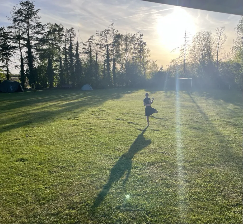 A faerie dancing barefoot in a field at golden hour, long shadow stretching across the grass, tents under the trees behind.